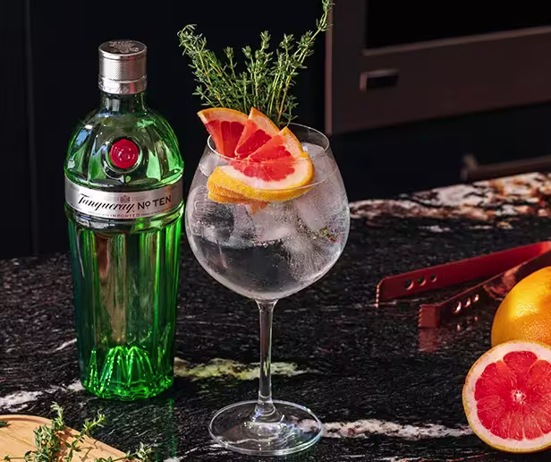A bottle of Tanqueray No. Ten gin next to a cocktail glass filled with ice, garnished with grapefruit slices and thyme, on a dark marble countertop with grapefruit halves nearby.