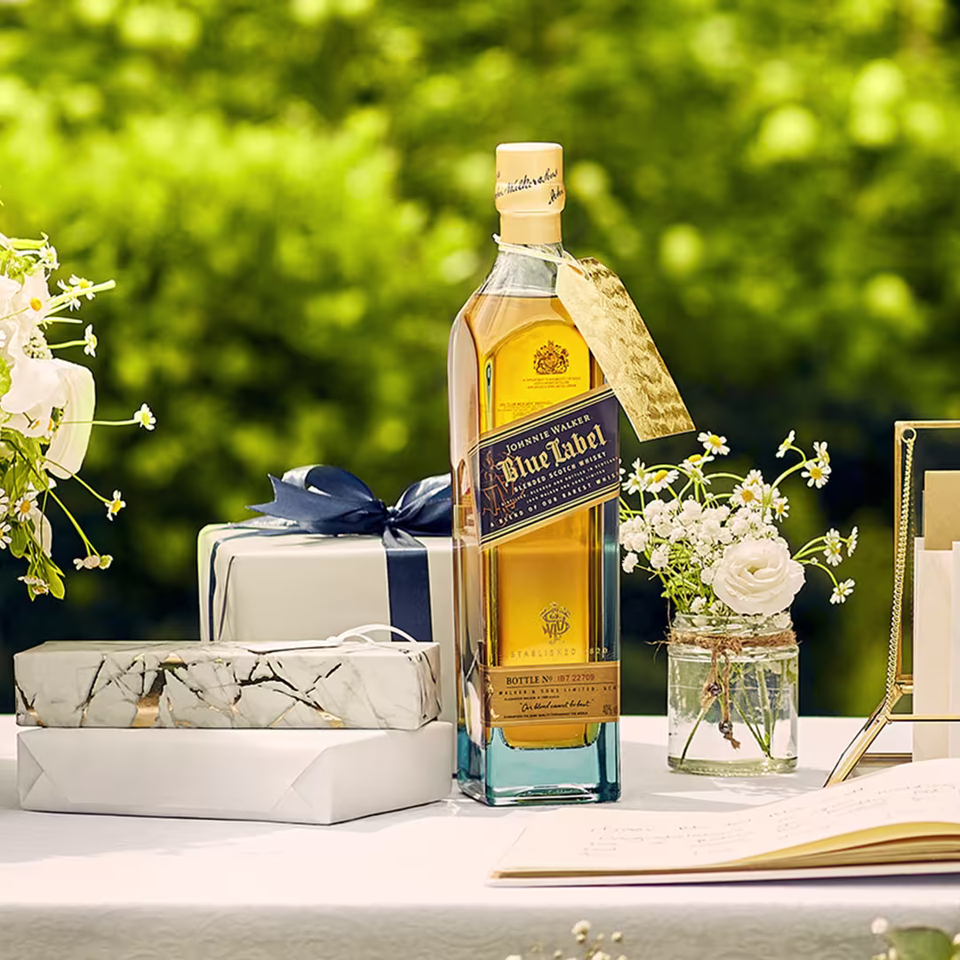 A bottle of Johnnie Walker Blue Label whisky is displayed on a table with wrapped gifts, flowers in a jar, and an open book, set outdoors with greenery in the background.