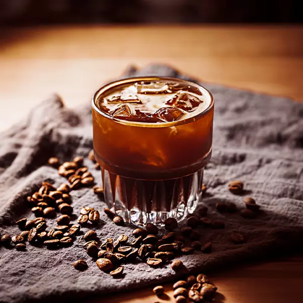 A glass of iced coffee sits on a textured cloth, surrounded by scattered coffee beans on a wooden surface.
