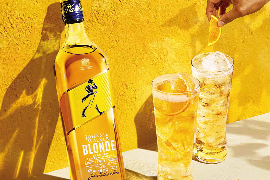 A bottle of Johnnie Walker Blonde whisky stands beside two tall glasses of whisky with ice and lemon garnish, against a bright yellow background.