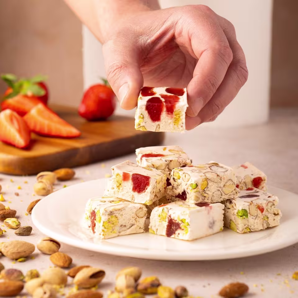 Cuadrados de turrón blando con pistachos y trozos de fresa confitada sobre un plato blanco, rodeados de frutos secos, con una mano destacando una pieza en primer plano.