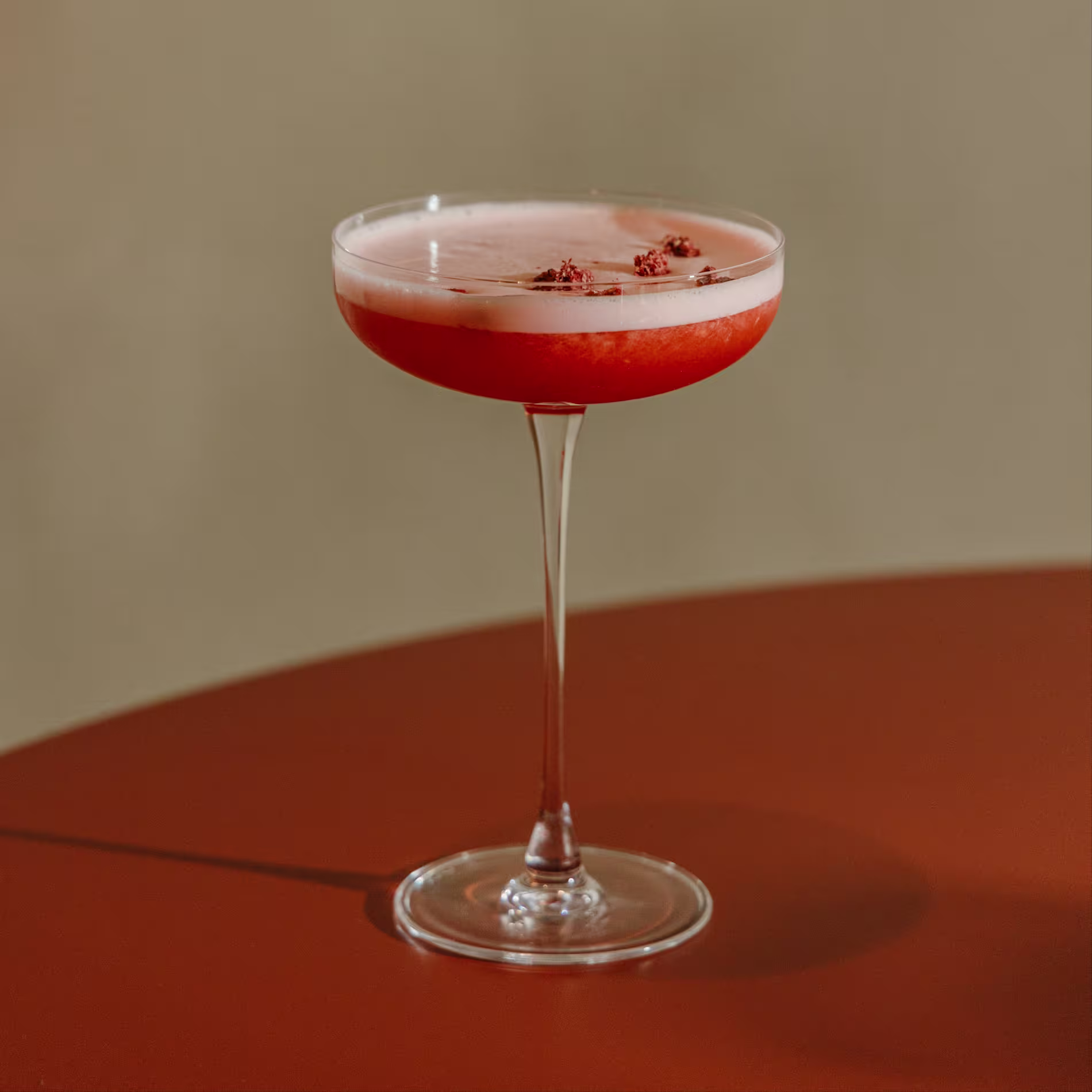 La imagen muestra una copa de cóctel con un líquido de color rosado con una capa espumosa en la parte superior. La bebida está decorada con pequeñas especias o hierbas marrones espolvoreadas encima.