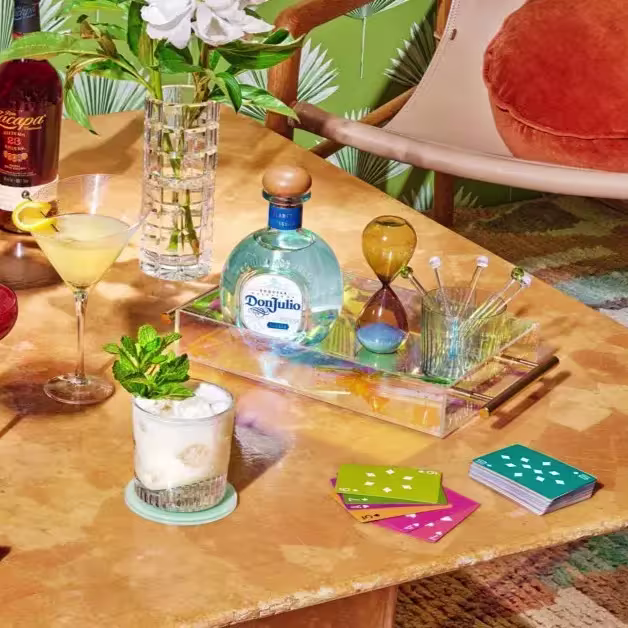 A marble table holds drinks, a bottle of Don Julio tequila, an hourglass, cocktail stirrers, playing cards, and a vase with white flowers. A chair with an orange cushion is beside the table.