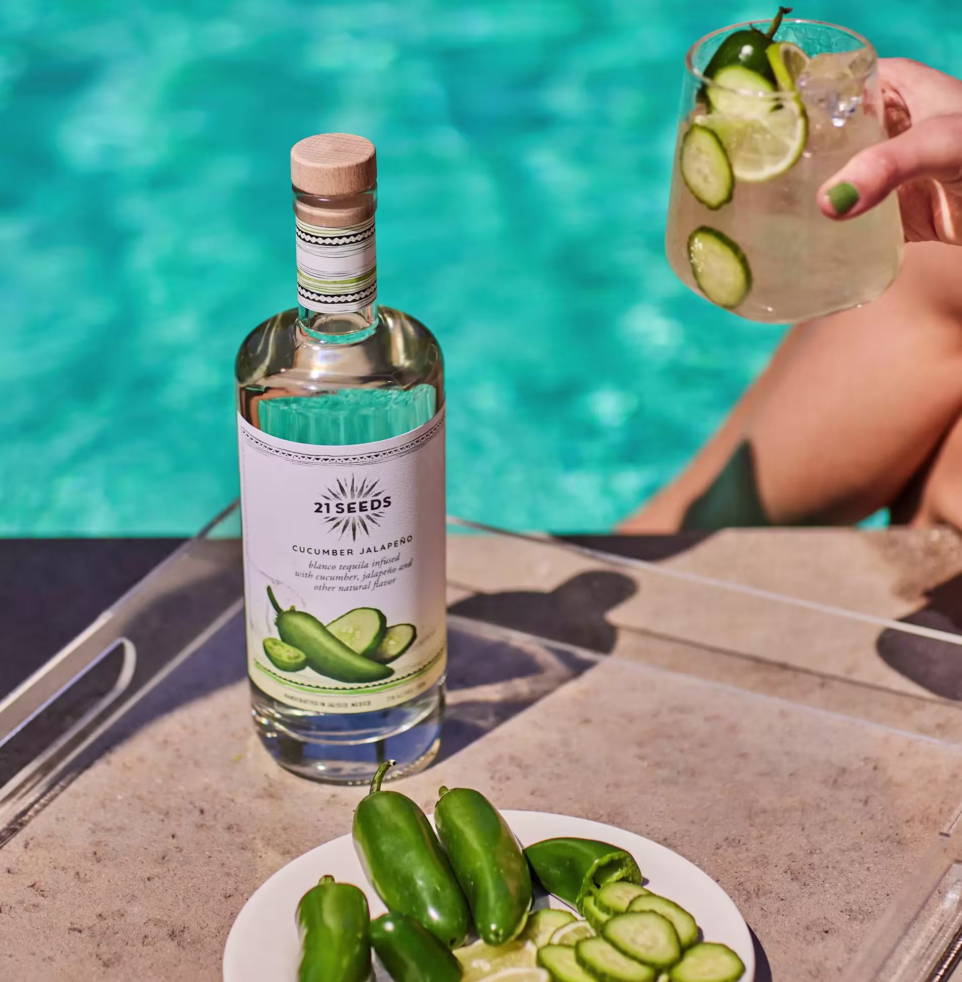 A bottle of 21 Seeds Cucumber Jalapeño tequila, a plate with sliced cucumbers and jalapeños, and a cocktail by a poolside.