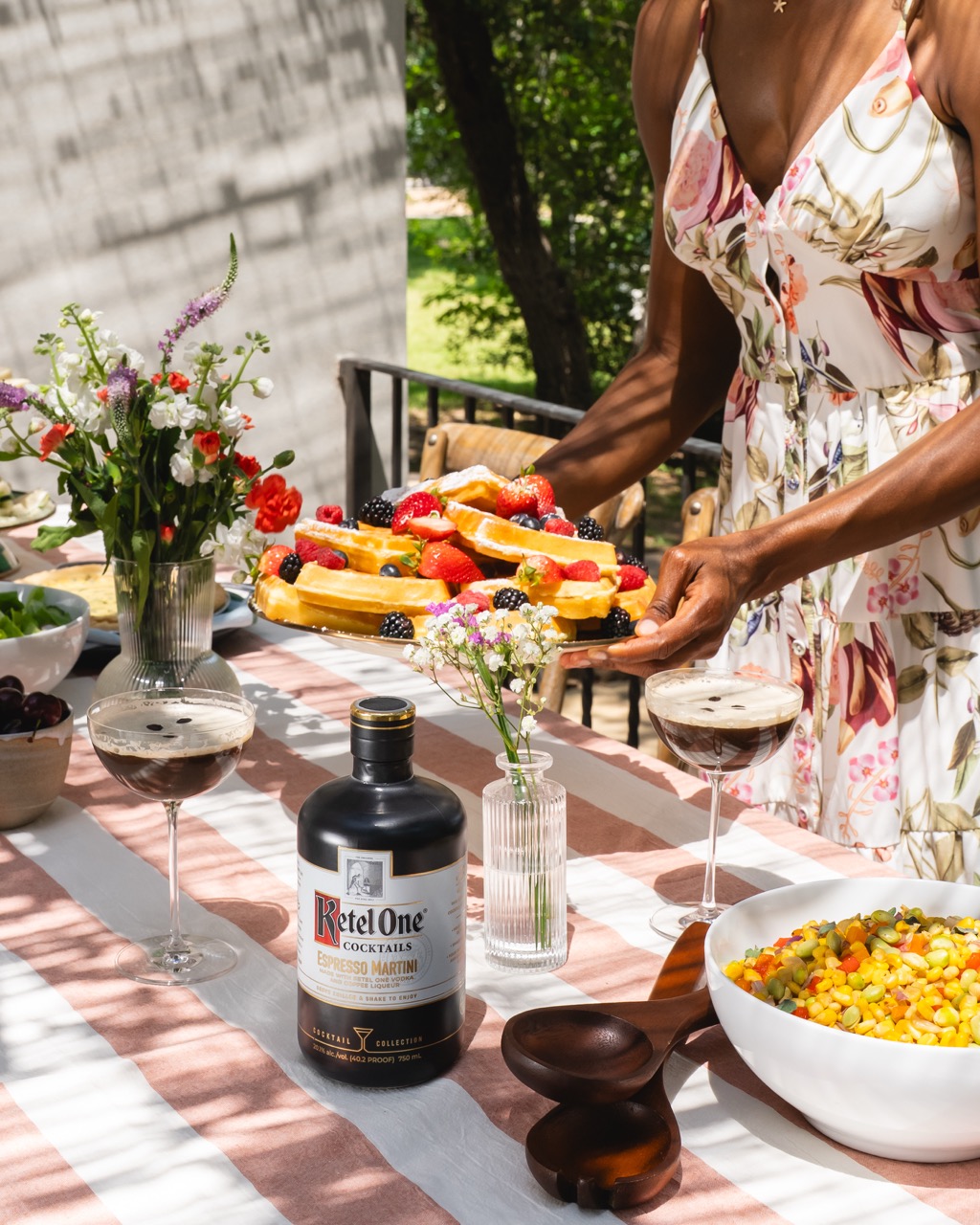 A woman in a floral dress sets a platter of fruit-topped pastries on a table with cocktails, a bottle of Ketel One Espresso Martini, flowers, and a bowl of corn salad.