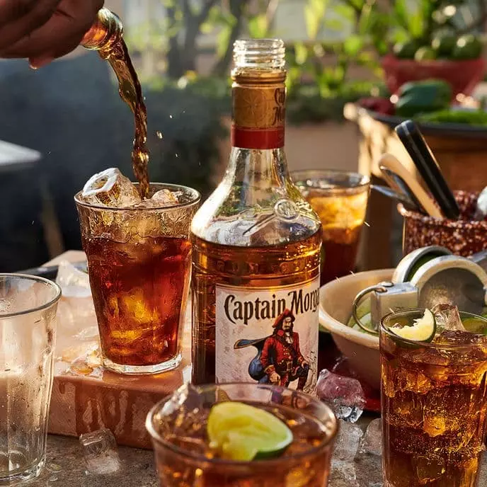 A bottle of Captain Morgan rum on a table with several glasses of iced drinks, lime wedges, and scattered ice cubes. A hand pours a dark liquid into one glass.
