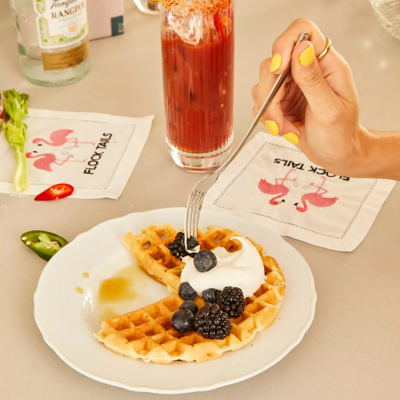A hand with yellow nail polish holds a fork over waffles topped with whipped cream, blackberries, and blueberries on a white plate, with a Bloody Mary and napkins nearby.