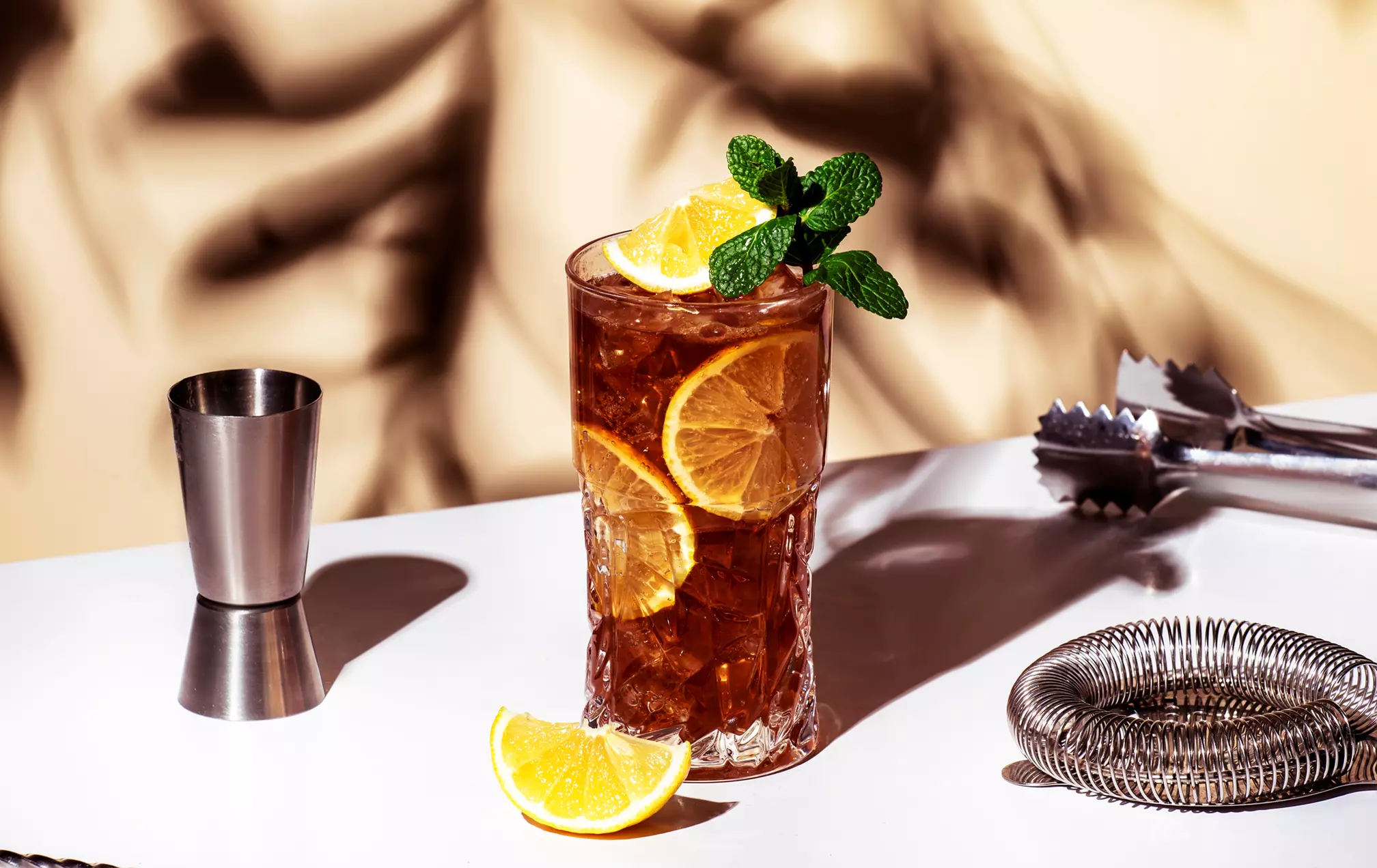 A glass of iced tea with lemon slices and mint garnish sits on a white surface, next to a jigger, cocktail strainer, tongs, and a lemon wedge.
