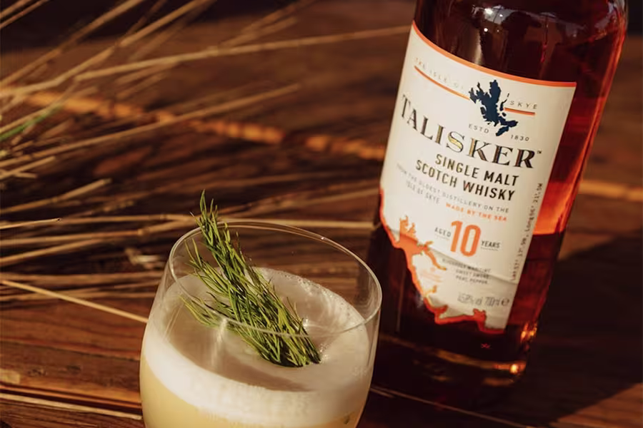 A glass of whisky cocktail garnished with herbs is placed beside a bottle of Talisker 10-Year-Old Single Malt Scotch Whisky on a wooden surface.