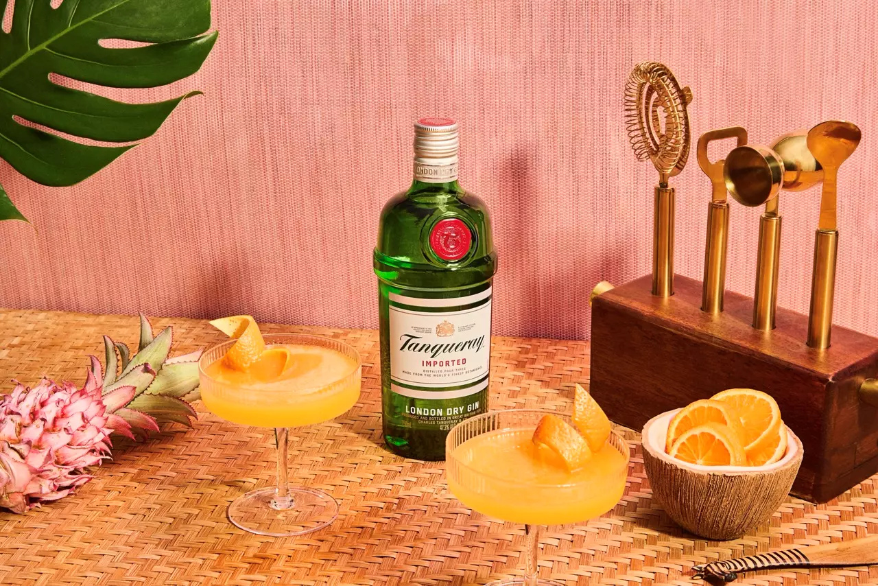 A bottle of Tanqueray gin is placed on a woven mat with two orange cocktails, a bowl of orange slices, a pineapple, and a decorative brass telephone against a pink background.