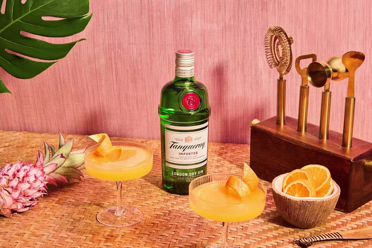 A bottle of Tanqueray gin is placed on a woven mat with two orange cocktails, a bowl of orange slices, a pineapple, and a decorative brass telephone against a pink background.