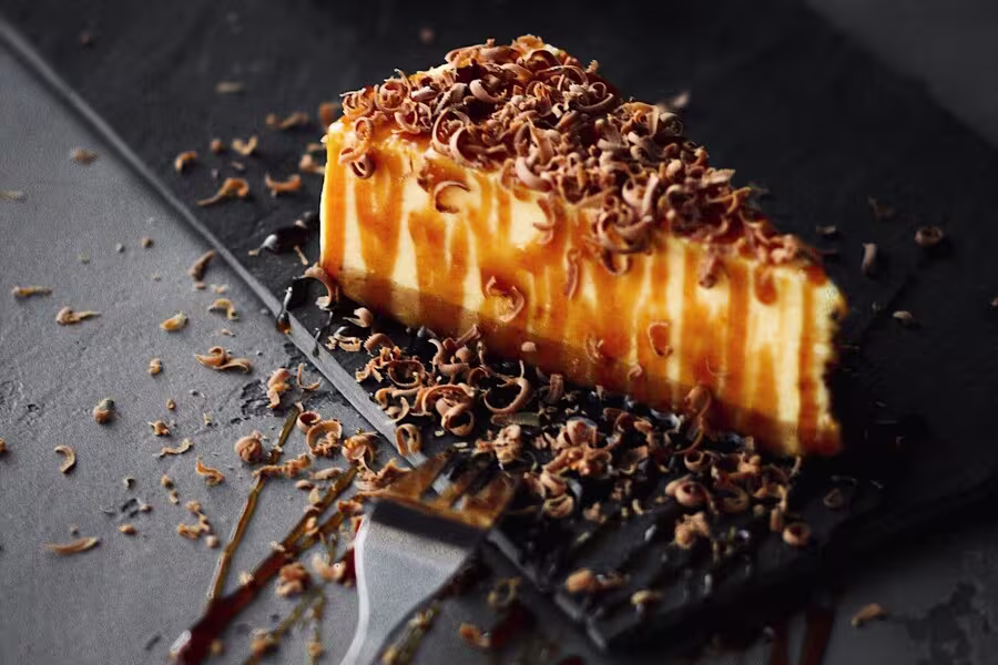 A slice of cheesecake topped with chocolate shavings and caramel sauce, served on a slate plate with a fork beside it.