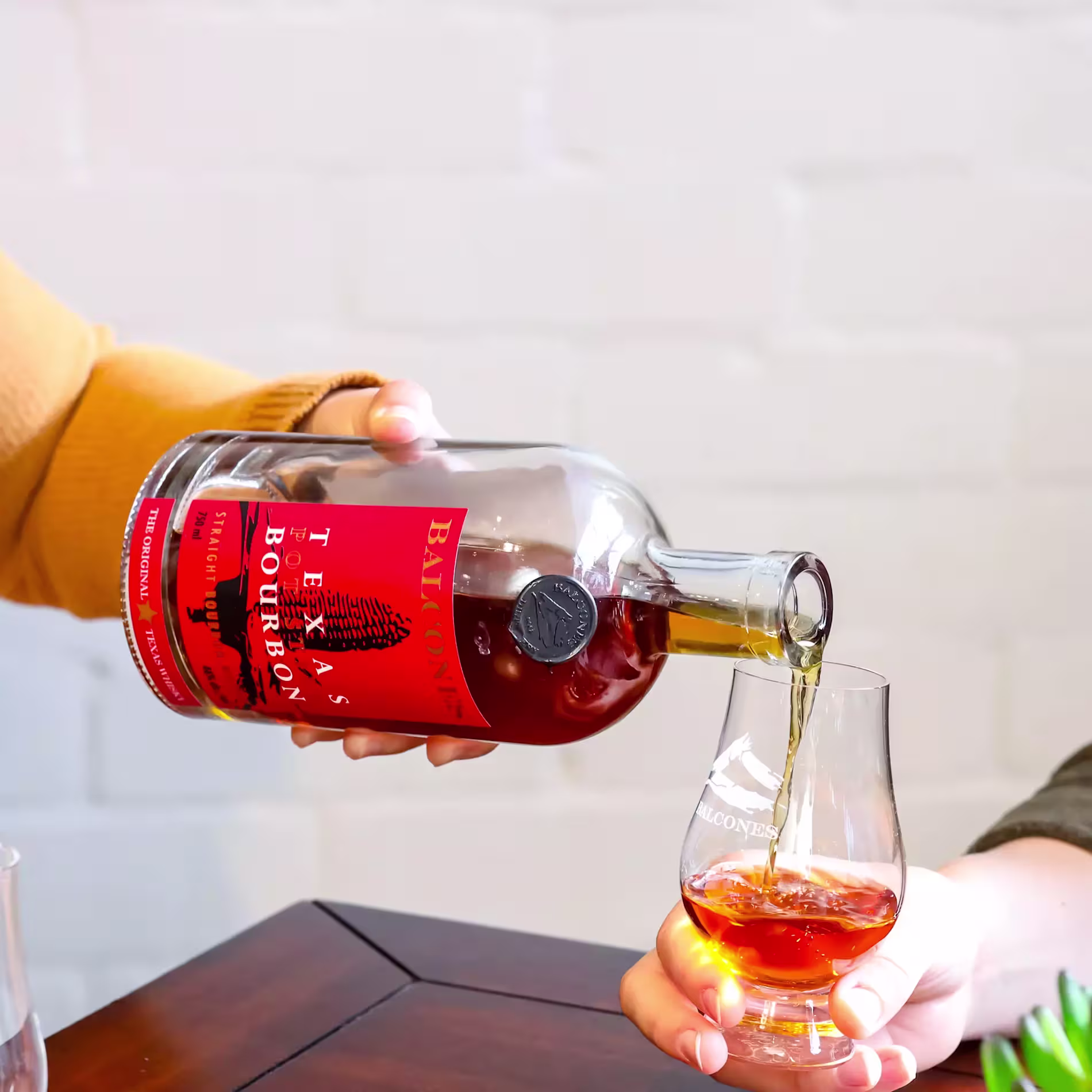 A person pours bourbon from a Balcones Texas Bourbon bottle into a whiskey glass held by another person on a wooden table.