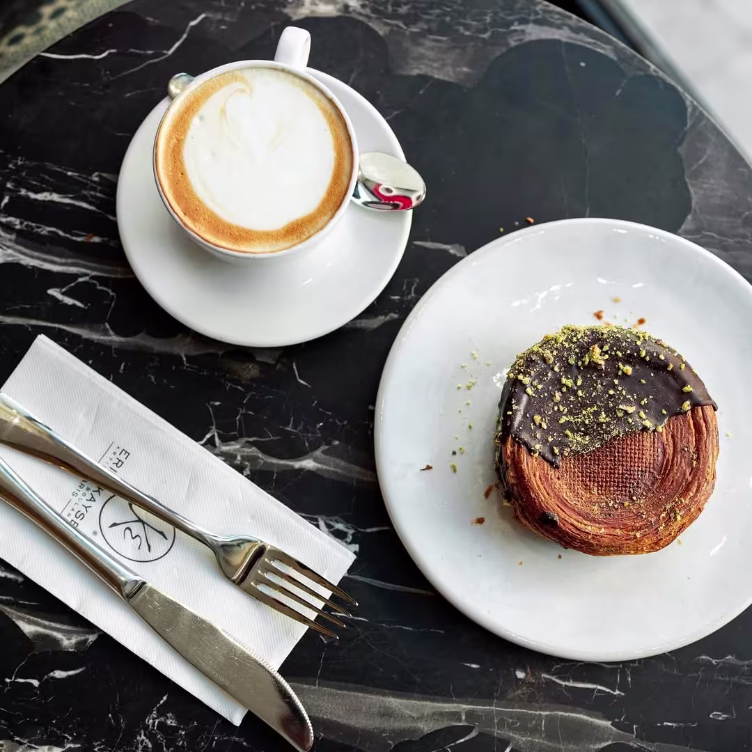 Café con espuma servido junto a un pastel hojaldrado con cobertura de chocolate y pistacho en una mesa de mármol negro en Maison Kayser.