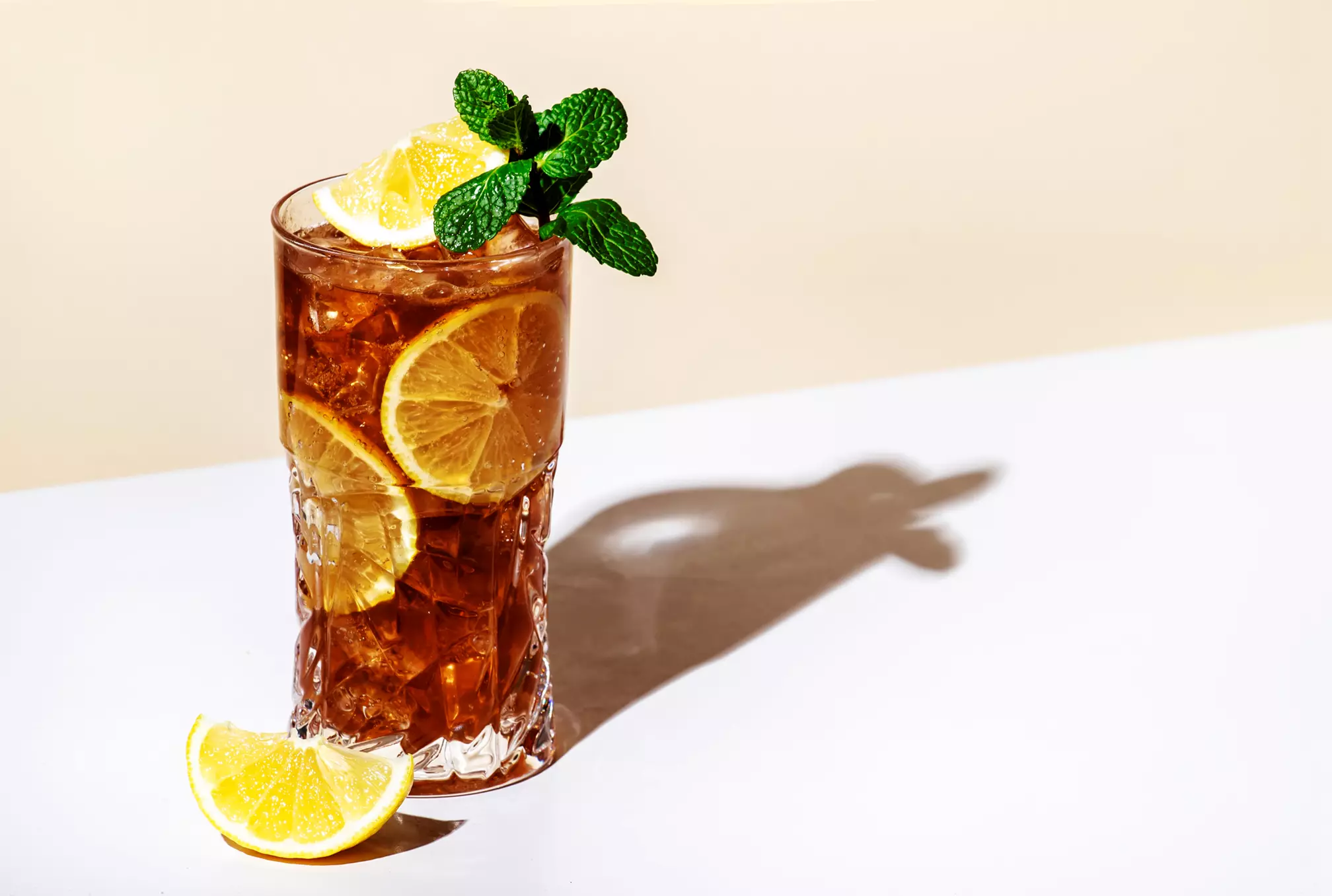 A glass of iced tea with lemon slices and mint garnish sits on a white surface, casting a shadow; a lemon wedge rests beside the glass.