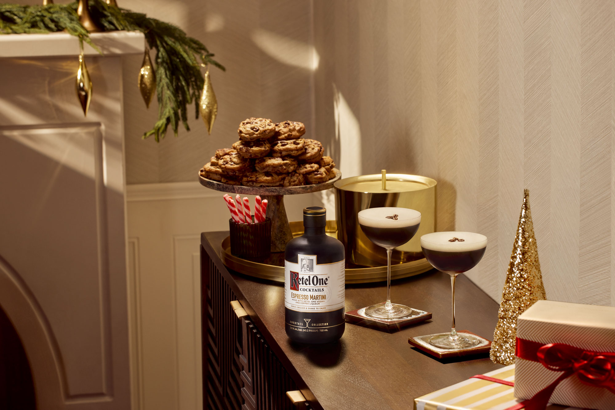 A bottle of Ketel One vodka, two espresso martinis, cookies on a stand, candy canes, a gold candle, and wrapped gifts on a decorated table near a fireplace.