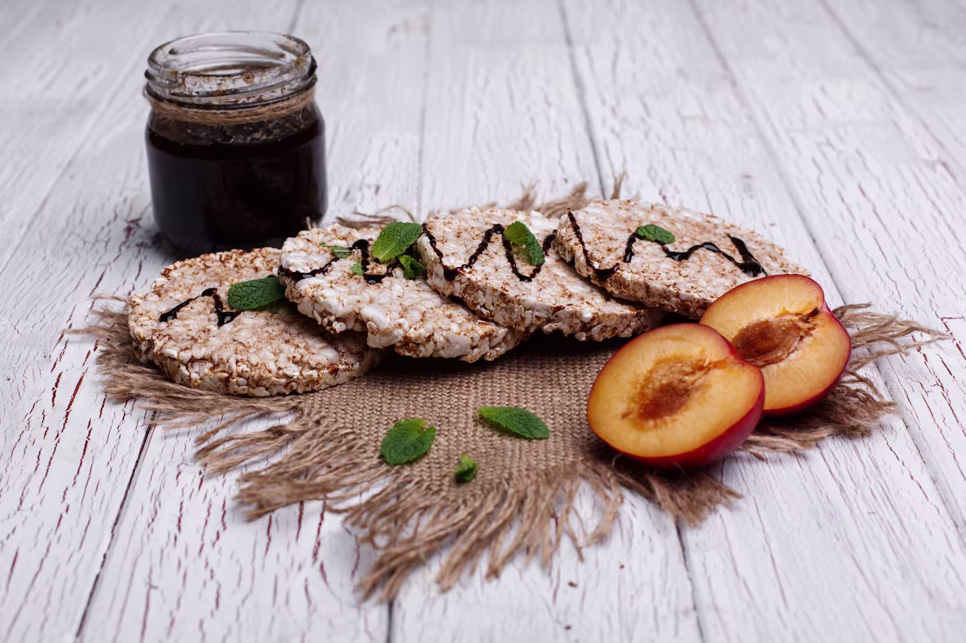 A jar of dark jam, rice cakes with chocolate drizzle, mint leaves, and a halved plum on a piece of burlap over a white wooden table.