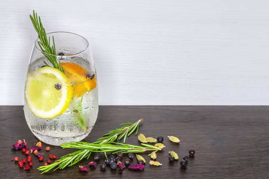 A glass of clear beverage with lemon, orange slices, and rosemary sprig, next to scattered spices and herbs on a dark wooden surface.