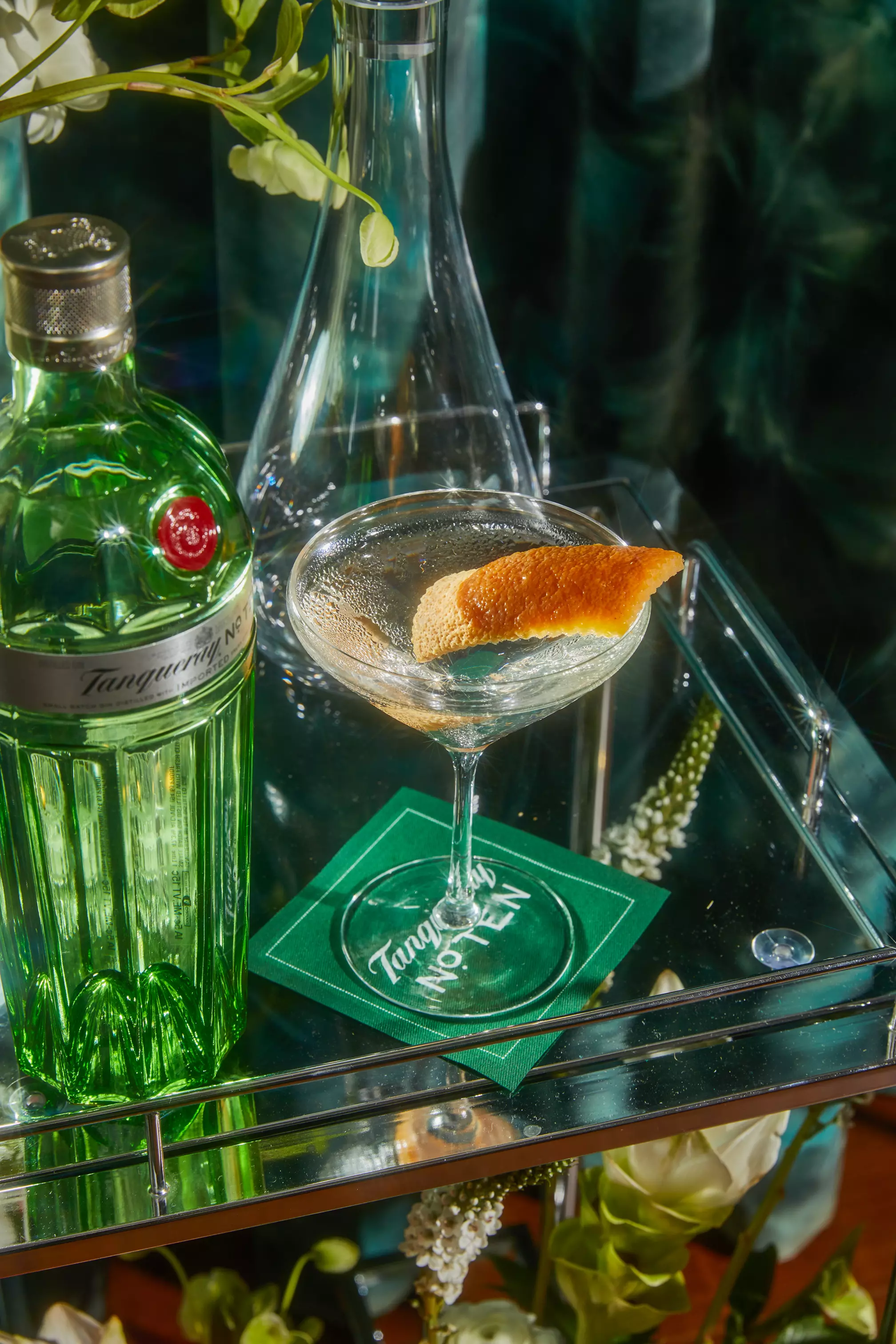 A bottle of Tanqueray No. Ten gin next to a martini glass with a citrus twist on a glass cart, with flowers and a green napkin reading Tanqueray No. Ten.