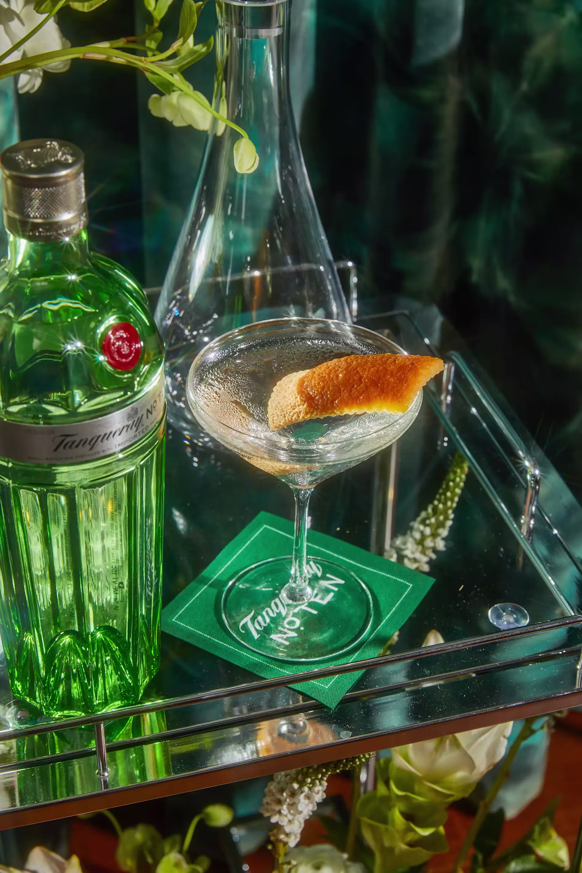 A bottle of Tanqueray No. Ten gin next to a martini glass with a citrus twist on a glass cart, with flowers and a green napkin reading Tanqueray No. Ten.