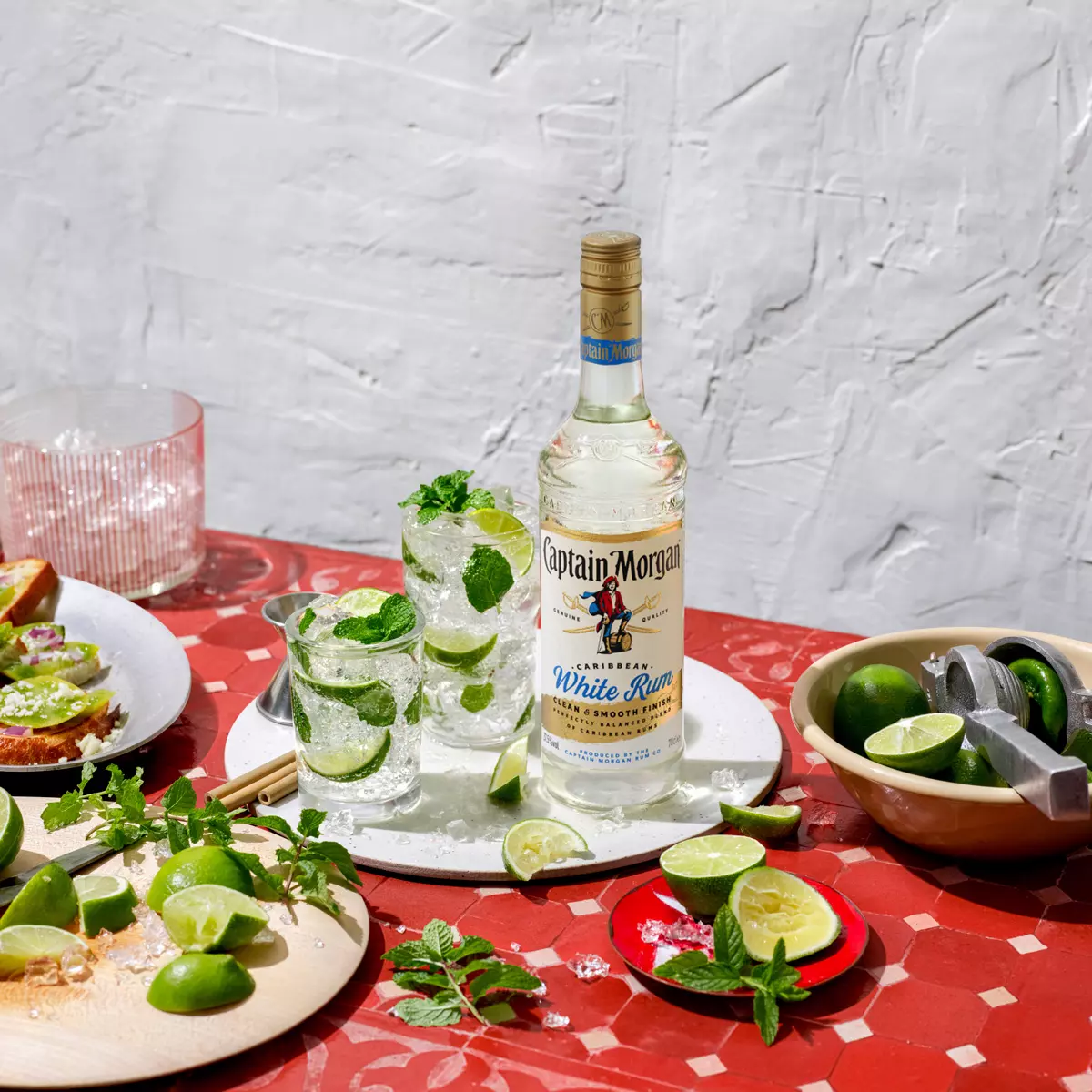 A bottle of Captain Morgan White Rum on a red table with two mojito cocktails, lime wedges, mint leaves, a cutting board, and a lime squeezer.