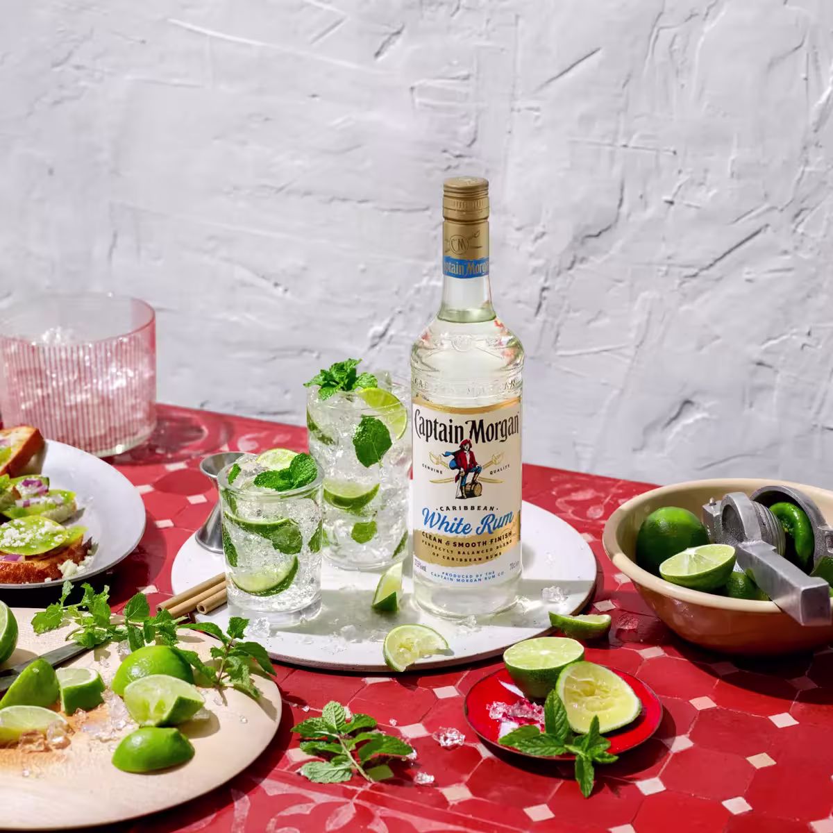 A bottle of Captain Morgan White Rum on a red table with two mojito cocktails, lime wedges, mint leaves, a cutting board, and a lime squeezer.