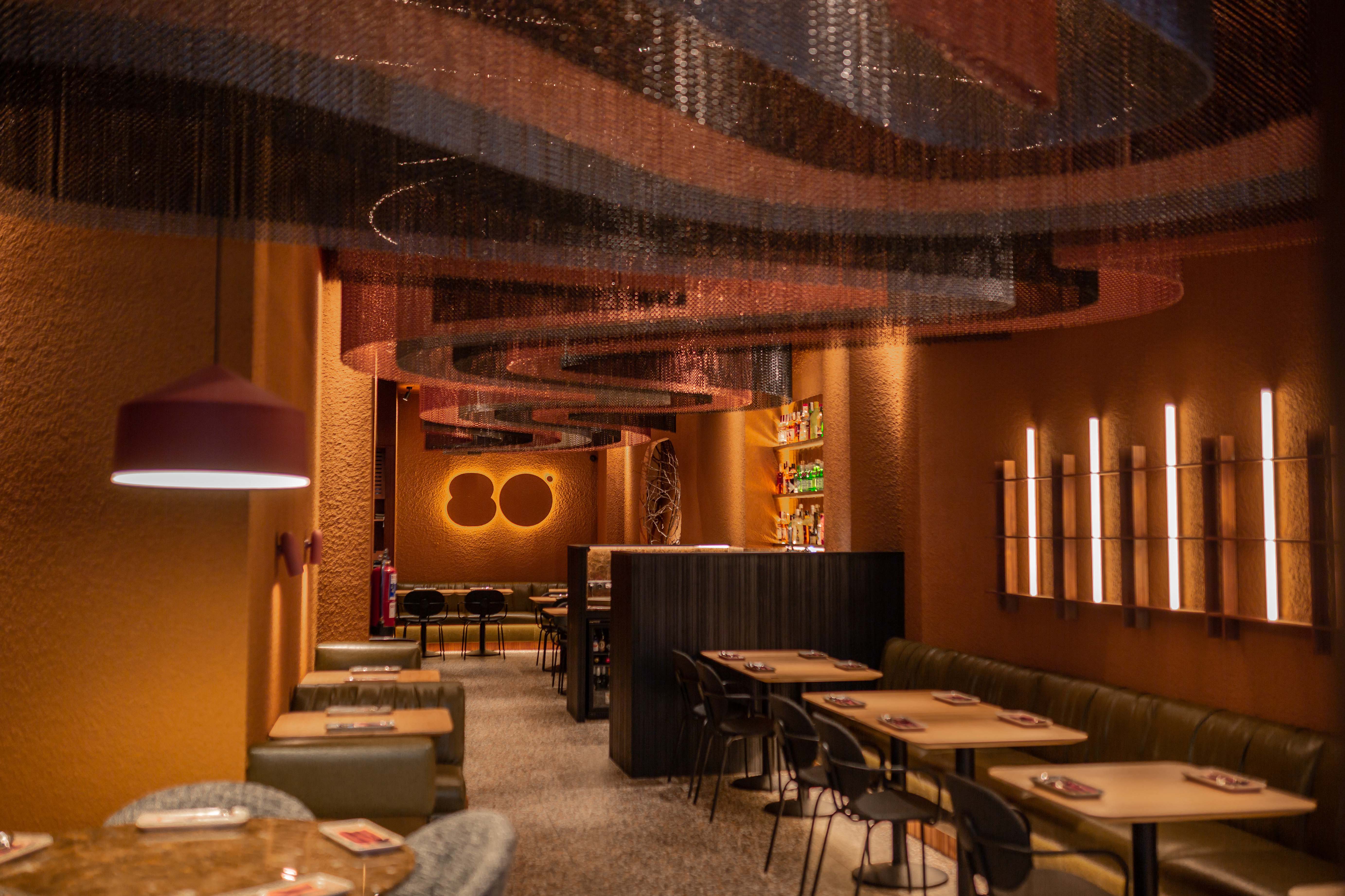 Interior de Ochenta Grados con luz cálida, decoración en tonos tierra, techo con formas onduladas y mesas dispuestas para disfrutar en un ambiente moderno.