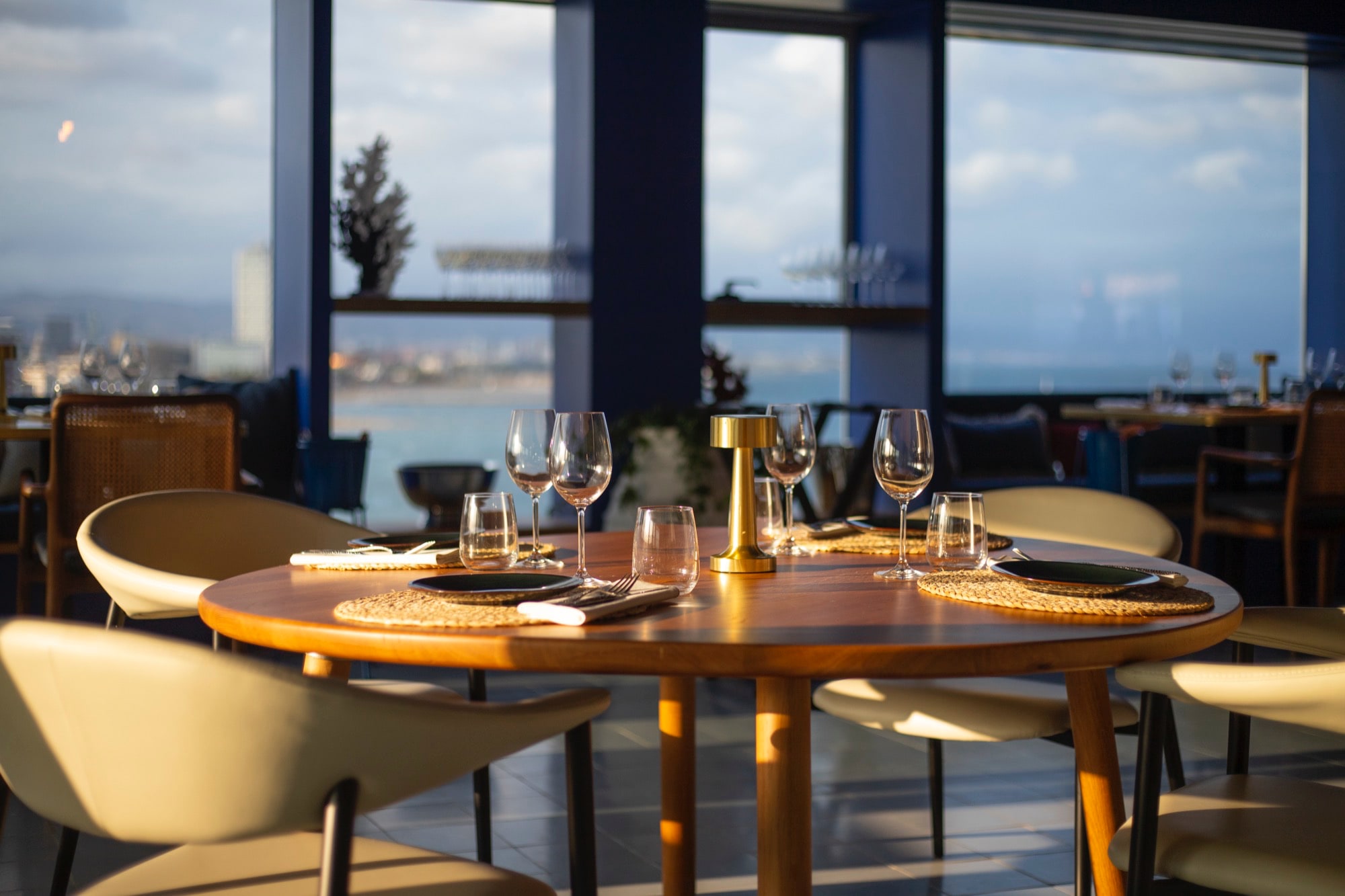 Azul Rooftop Barceloneta muestra una mesa redonda de madera cuidadosamente decorada con copas de cristal, lámpara dorada y vistas panorámicas al mar.