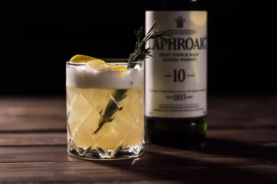 A glass of whisky sour garnished with a lemon peel and rosemary, placed in front of a bottle of Laphroaig 10 Year Old Scotch whisky on a wooden surface.