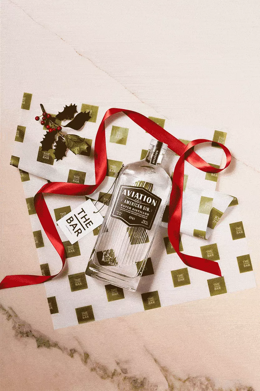 A bottle of Aviation American Gin with a red ribbon, holly sprig, and green-and-white patterned paper on a marble surface.