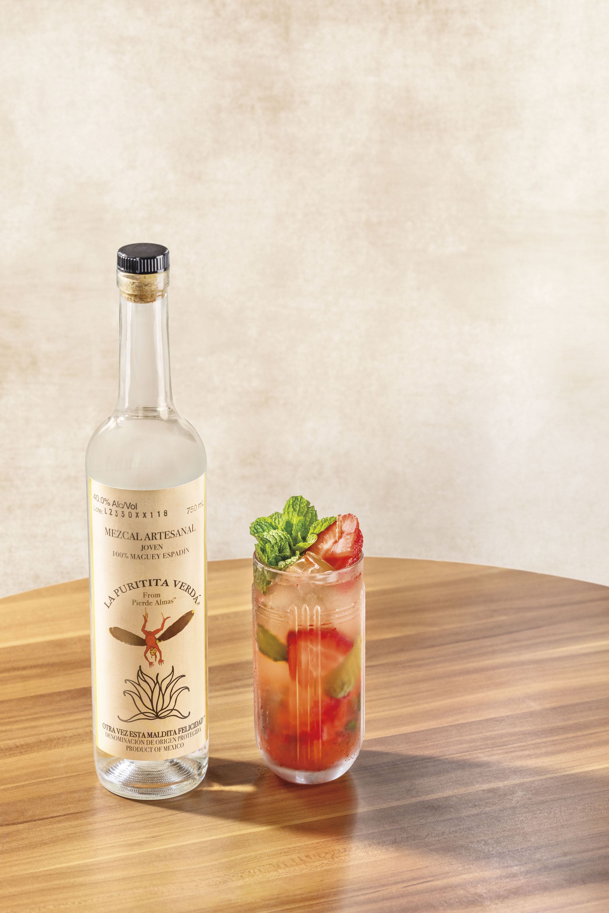A bottle of La Puritita Verda mezcal stands next to a glass cocktail garnished with mint and sliced strawberries on a wooden table.