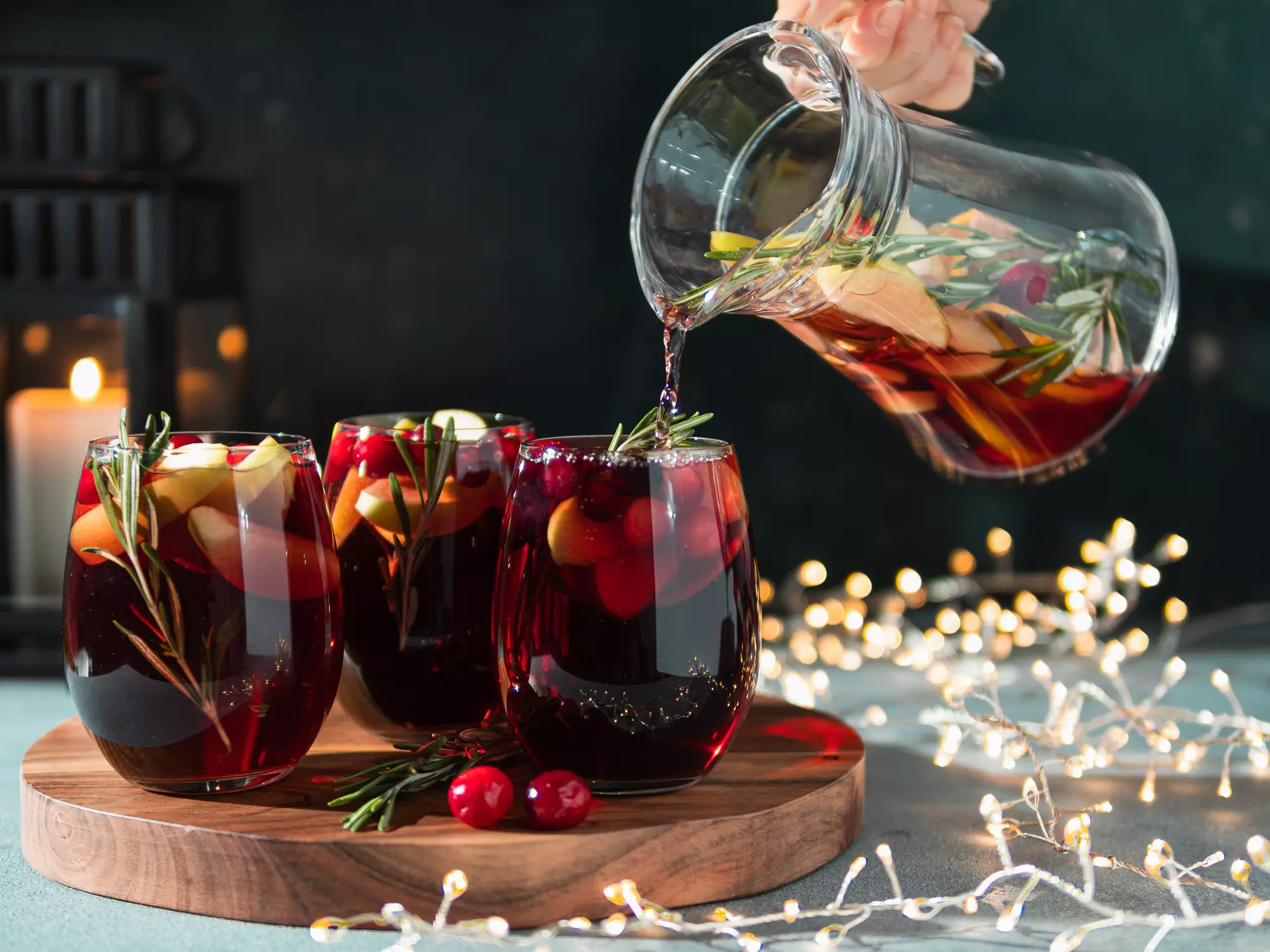 A hand pours red sangria with fruit and rosemary from a glass pitcher into stemless glasses on a wooden tray, with string lights and a lit candle in the background.
