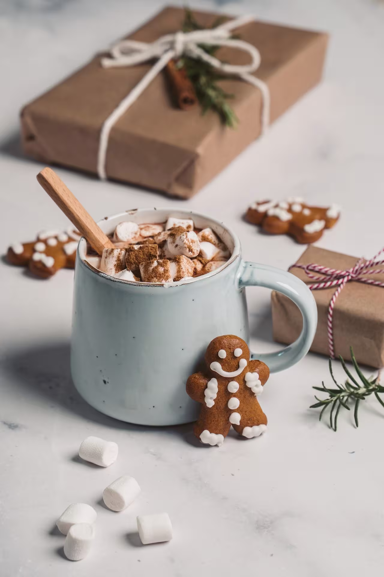 A blue mug filled with hot chocolate, marshmallows, and a wooden spoon sits on a table with gingerbread cookies, small presents, and scattered marshmallows.