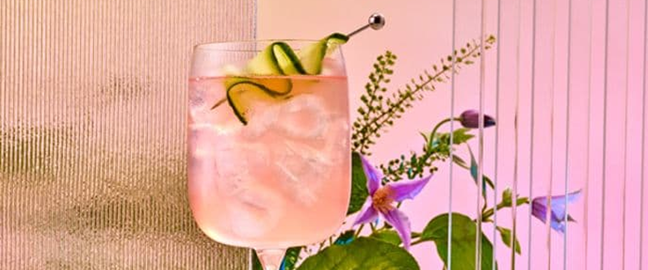 Cóctel de ginebra rosa con guarnición de cinta de pepino y fondo floral
