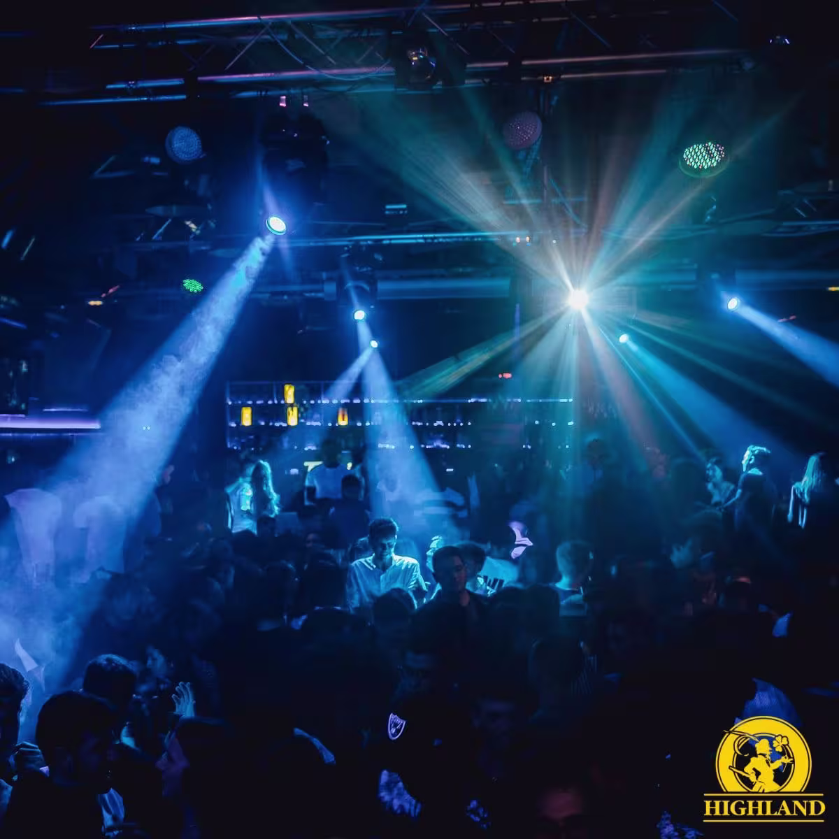 " Highland: Ambiente vibrante en la pista de baile, iluminada por luces azules y efectos de humo que crean una atmósfera enérgica. La multitud disfruta de la música en un entorno dinámico y lleno de vida."