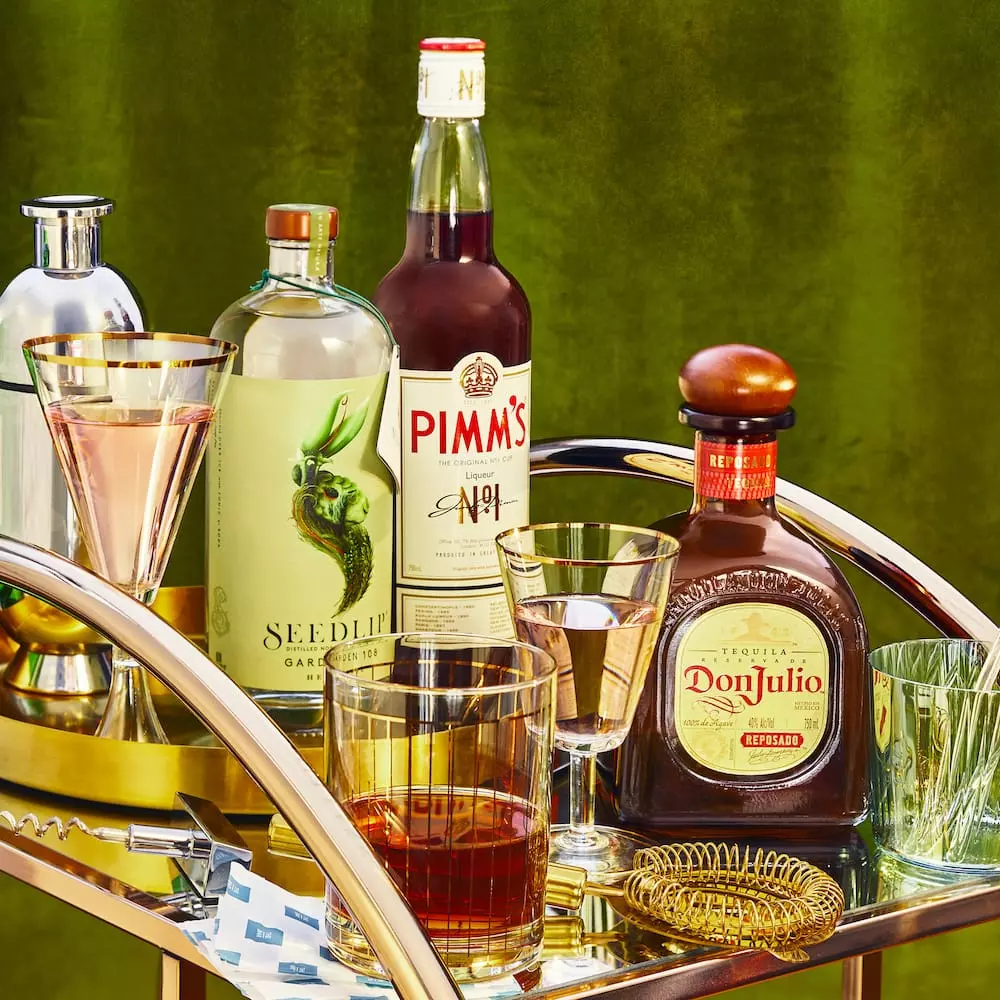 A bar cart with liquor bottles, including Seedlip, Pimm’s No.1, and Don Julio, along with cocktail glasses, a shaker, and a strainer on a gold tray against a green background.