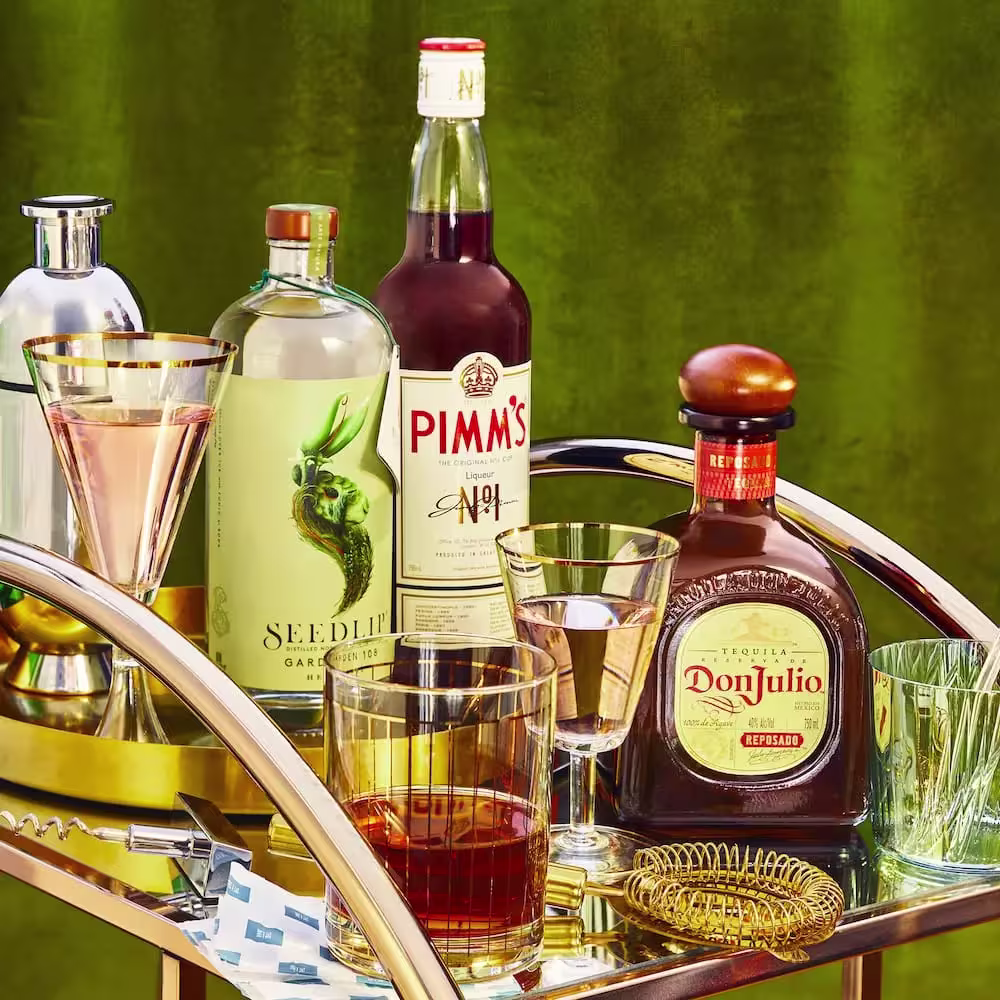 A bar cart with liquor bottles, including Seedlip, Pimm’s No.1, and Don Julio, along with cocktail glasses, a shaker, and a strainer on a gold tray against a green background.