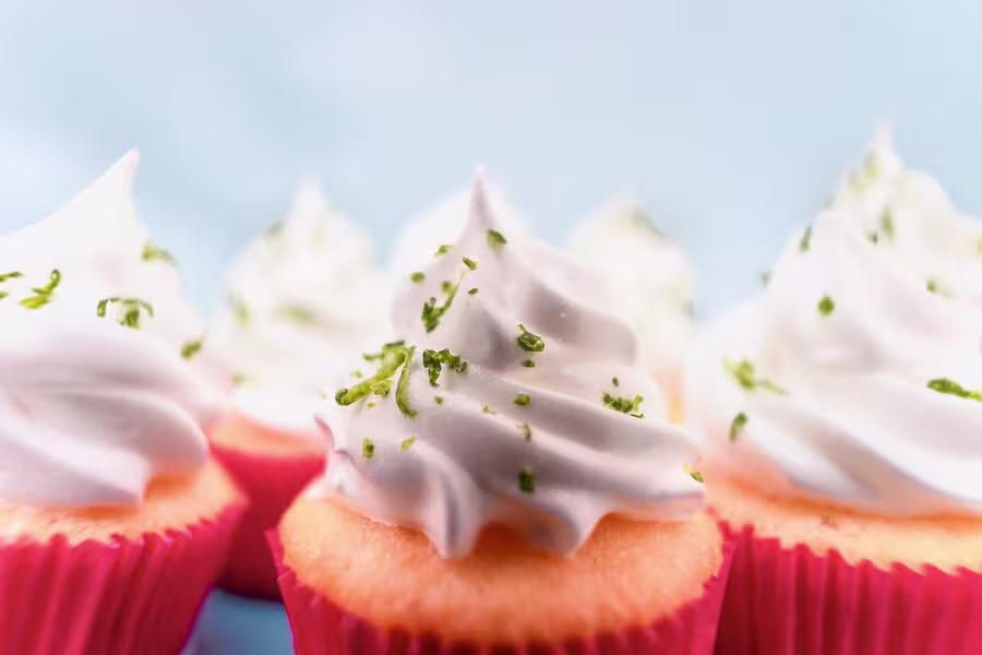 Close-up of several cupcakes with light orange bases in pink wrappers, topped with swirls of white frosting and sprinkled with green zest.