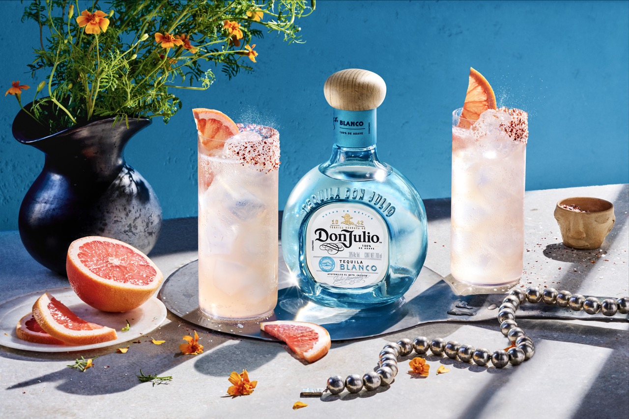 A bottle of Don Julio Blanco tequila with two grapefruit cocktails, garnished with grapefruit slices and chili, sits on a table with flowers, beads, and a cut grapefruit.