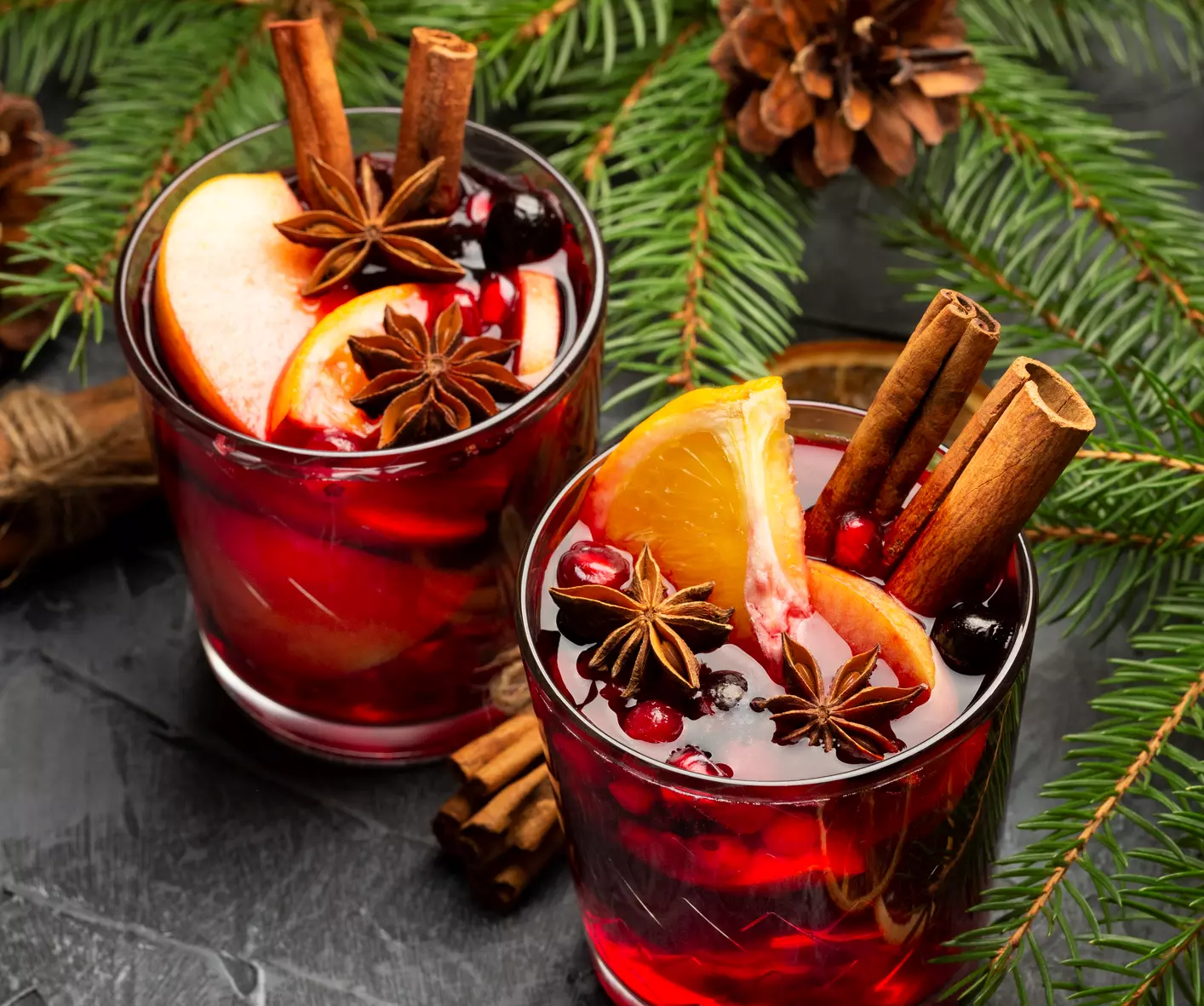 Two glasses of mulled wine garnished with orange slices, apple slices, cinnamon sticks, star anise, cranberries, and surrounded by pine branches and pine cones.