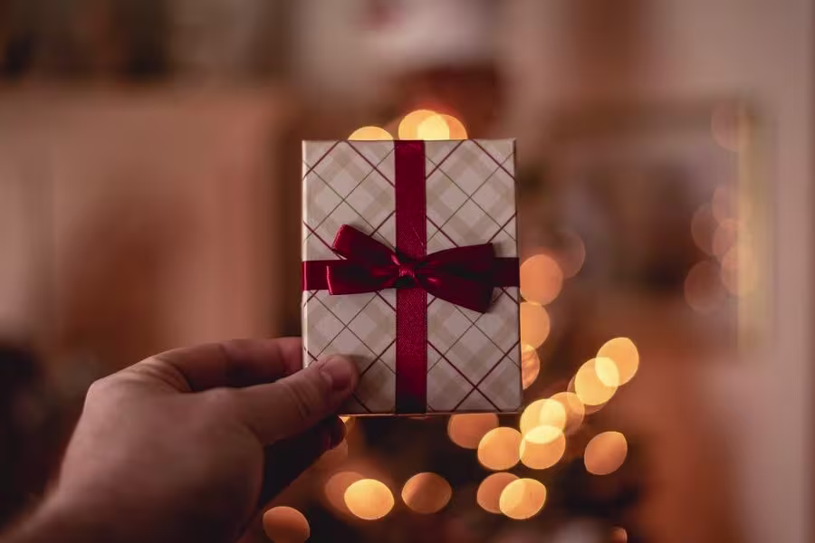 A hand holds a small, wrapped gift box with a red ribbon and bow. Warm, blurred lights are in the background.