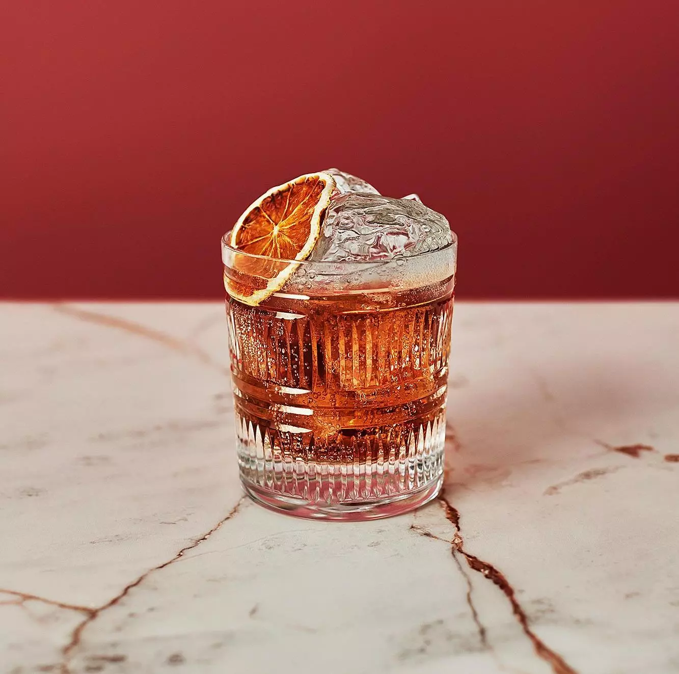 A glass of iced cocktail garnished with a dried orange slice sits on a white marble surface against a red background.