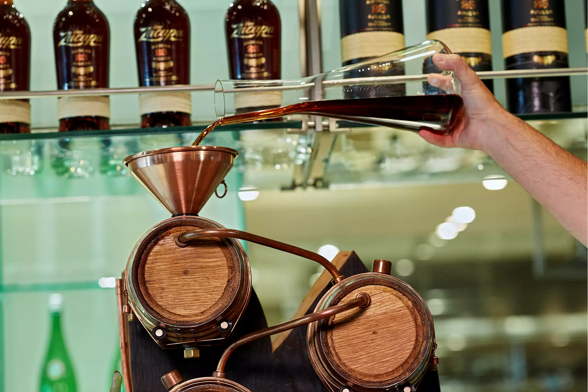 A hand pours dark liquid from a glass vessel into a copper funnel leading to wooden barrels; bottles are displayed on a shelf in the background.