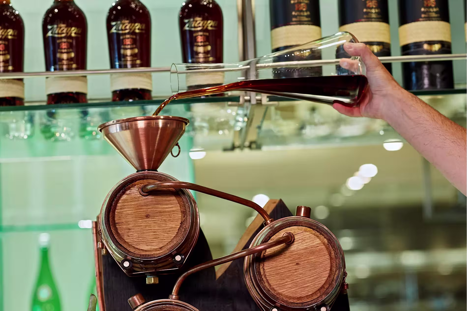 A hand pours dark liquid from a glass vessel into a copper funnel leading to wooden barrels; bottles are displayed on a shelf in the background.