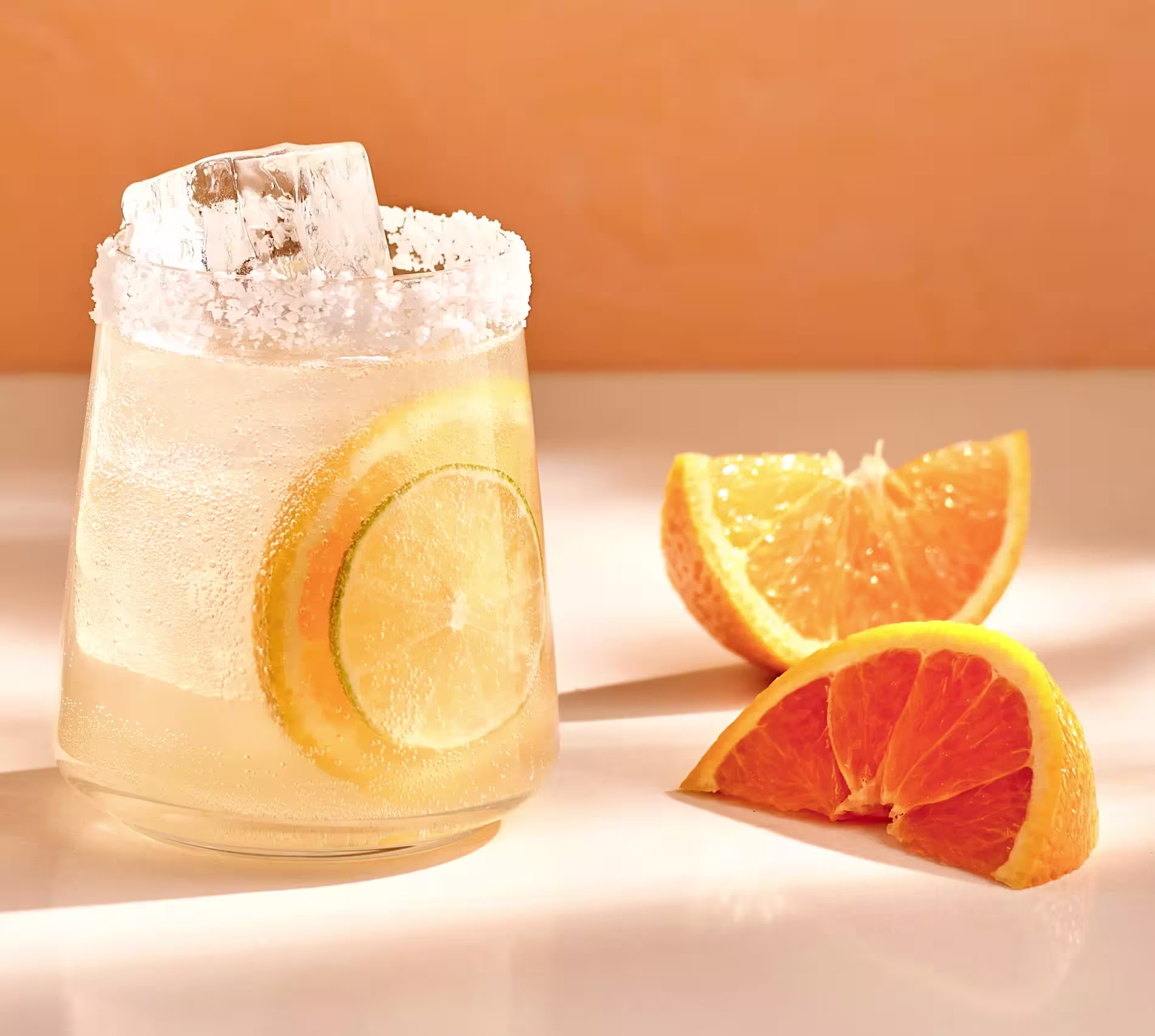 A cocktail with lemon and lime slices, ice, and a salted rim sits next to three orange wedges on a white surface with a warm background.