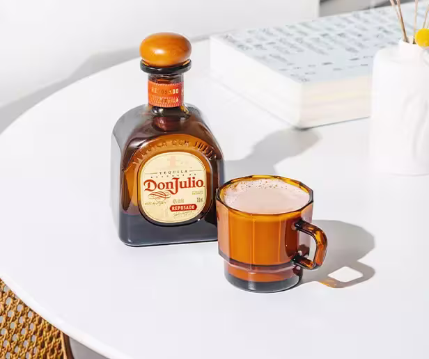 A bottle of Don Julio tequila sits next to a brown glass cup filled with a foamy beverage on a white table.
