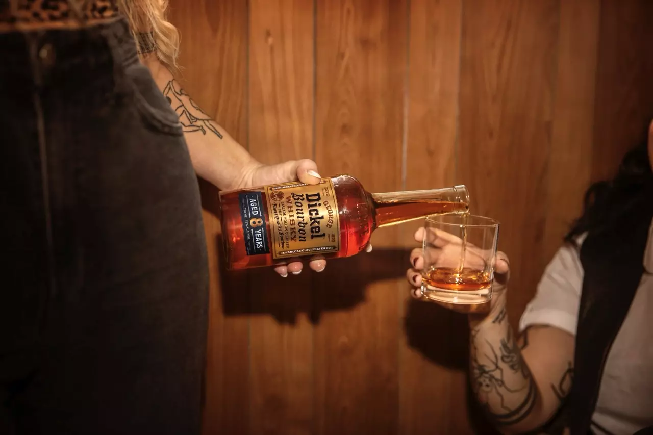 A person pours Dickel Bourbon whiskey from a bottle into a glass held by another person against a wooden wall background.