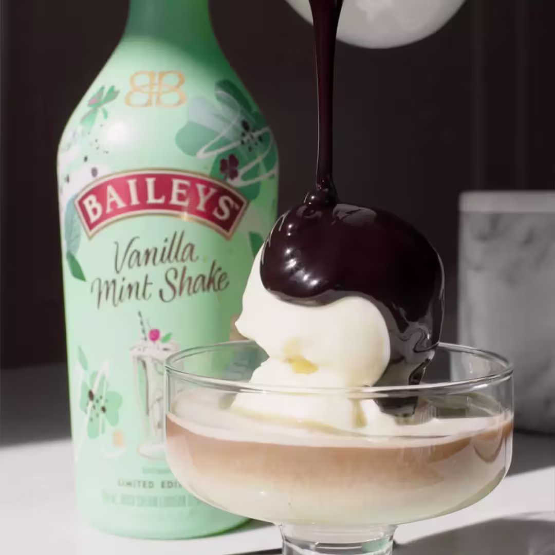 A bottle of Baileys Vanilla Mint Shake is behind a coupe glass with vanilla ice cream, over which chocolate sauce is being poured.