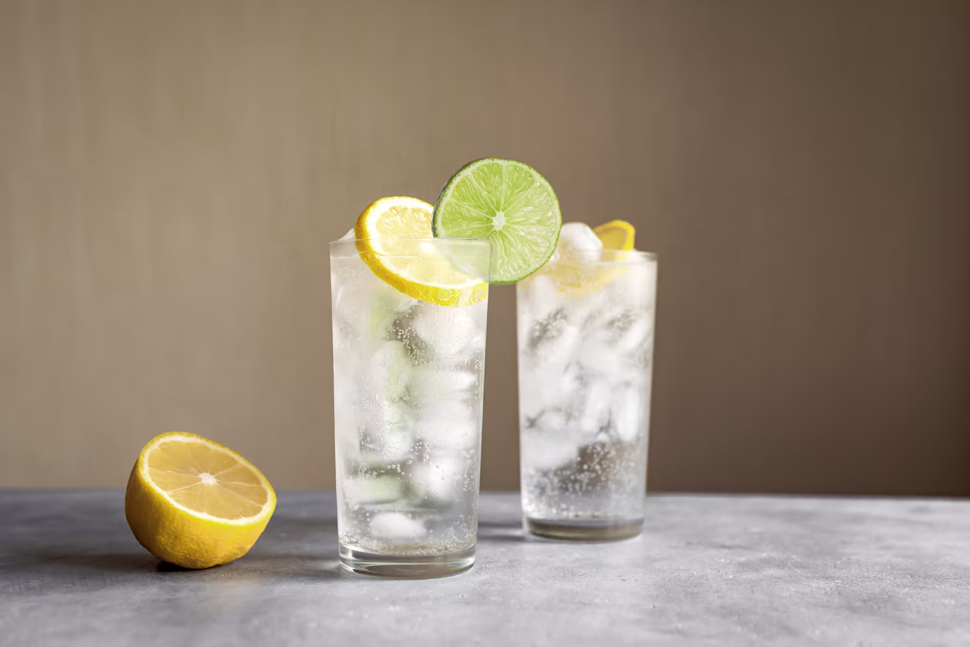 Two tall glasses of gin cocktails filled with ice and garnished with lemon and lime slices, on a grey stone surface.