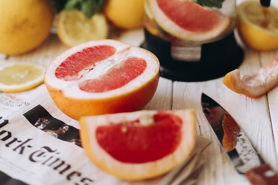 A halved grapefruit and a grapefruit wedge sit on a newspaper next to a knife, with lemon slices and a fruit juicer in the background.